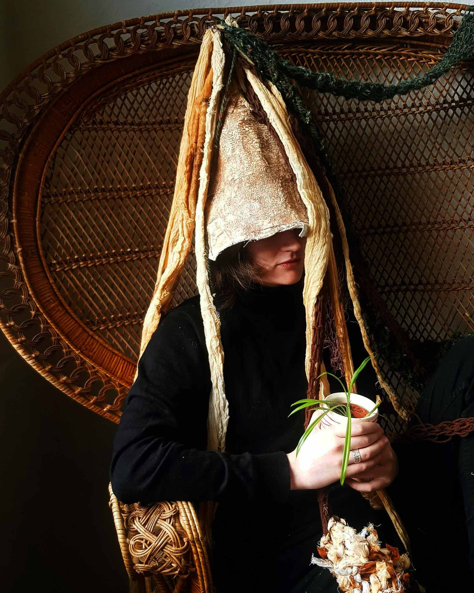 Image of a person wearing a gnome like hat with kitted tendrils, holding a spider plant while sat in a wicker chair