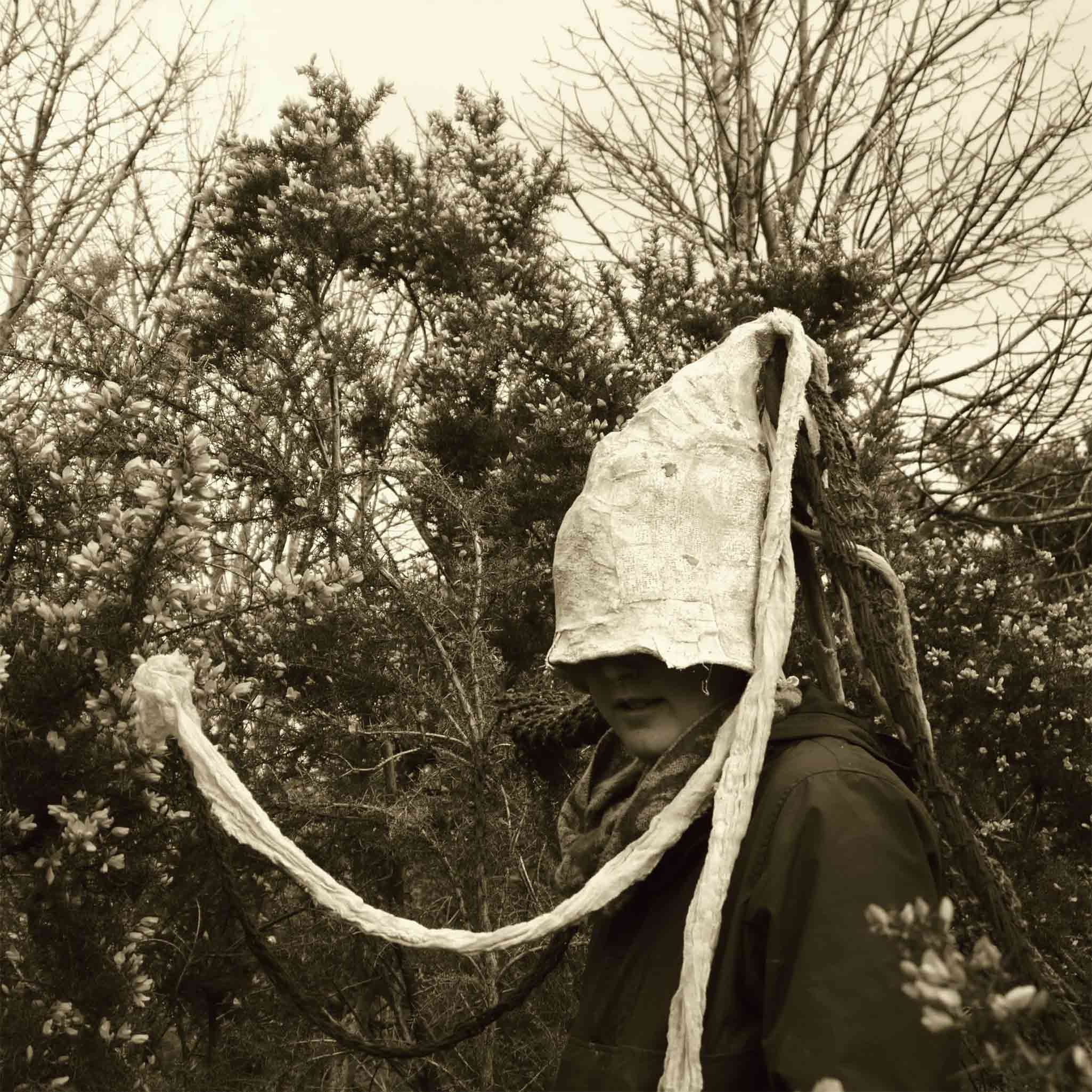 Black and white image with a person wearing a gnome like hat with tendrils surrounded by gorse bushes.