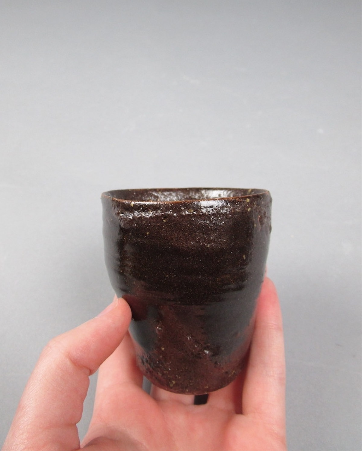 A small cup glazed in dark brown and green, held in the artist's hand.