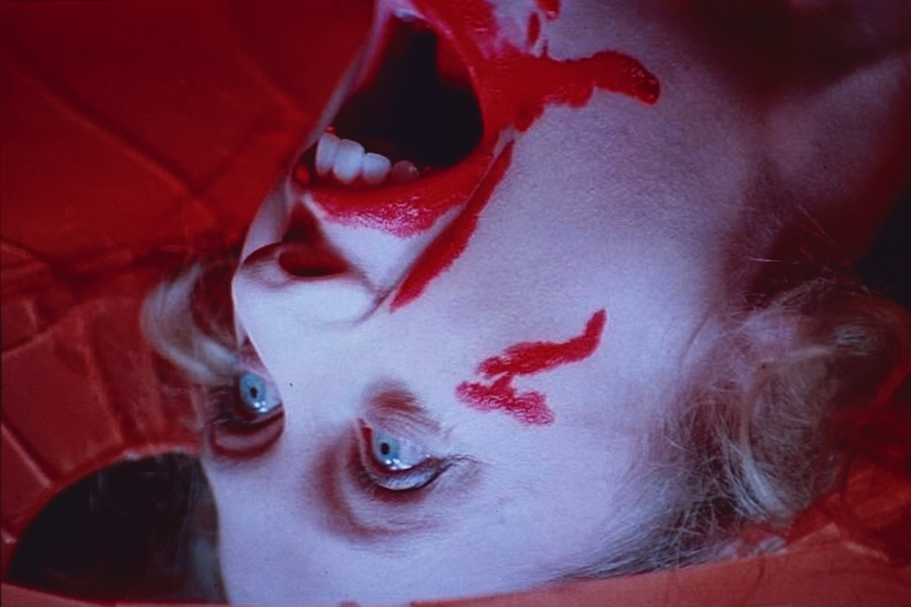 A still from Suspiria, a woman throws her head backwards, the screen drenched in kitsch red blood