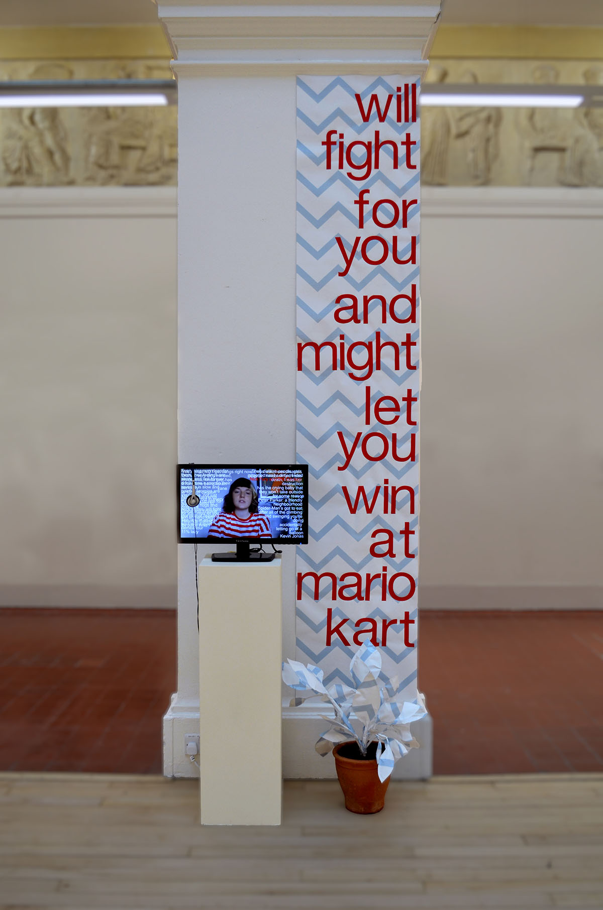 Installation shot consisting of wallpaper with text on pillar, monitor on plinth and a wallpaper potted plant