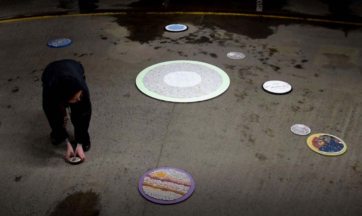 Person in all black crouching down to mosaic. In the background and foreground there are 13 mosaics depicting the solar system