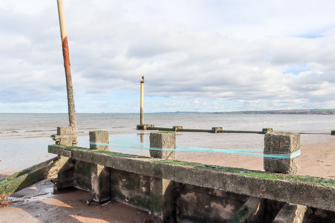 Strands of blue yarn wound around three groynes on a beach. The thread around two of the groynes has been crossed over, forming an 'x' shape. 