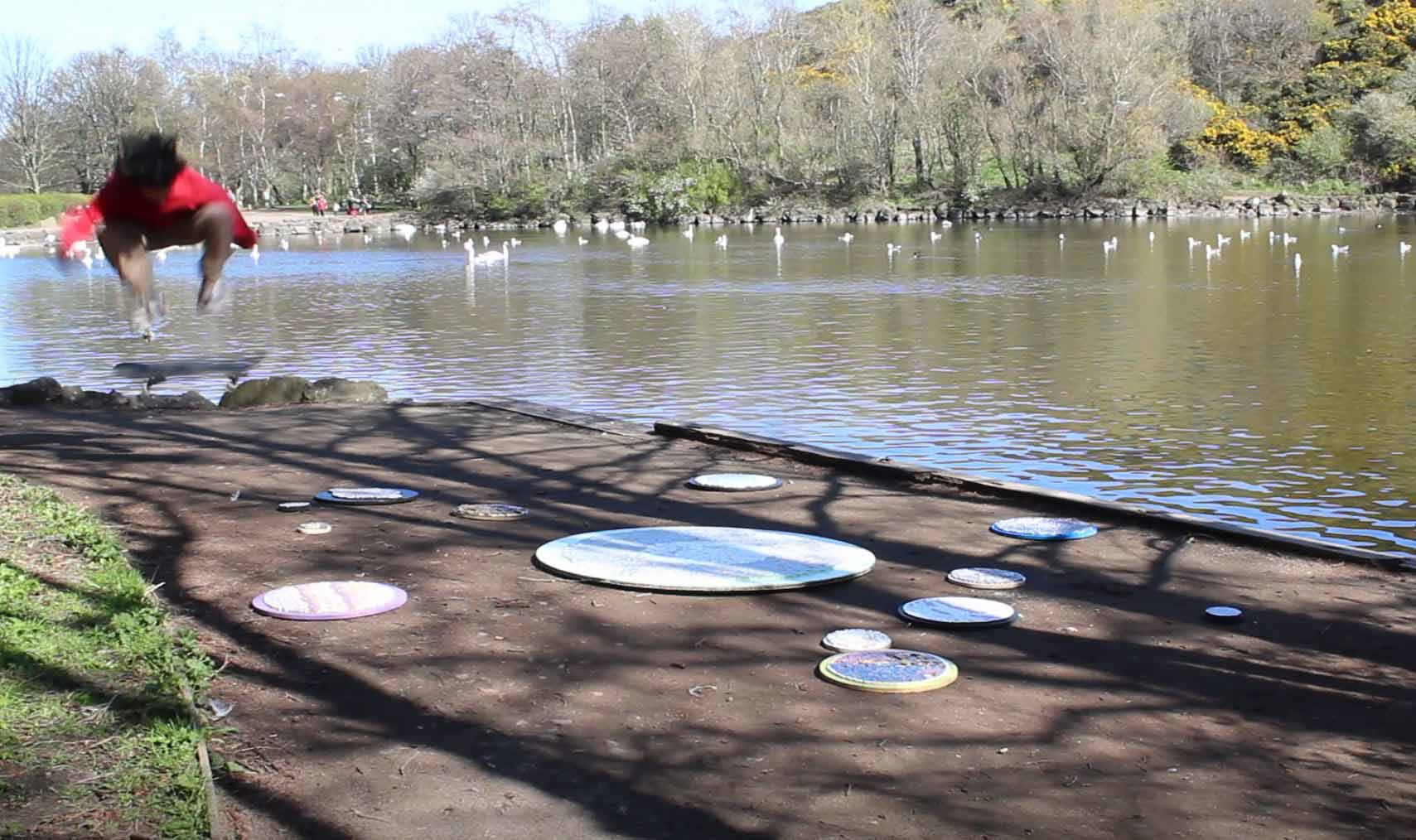 Man jumping off a skateboard and forming lotus position. In the background there is a lake with swans and in the foreground there are mosaics
