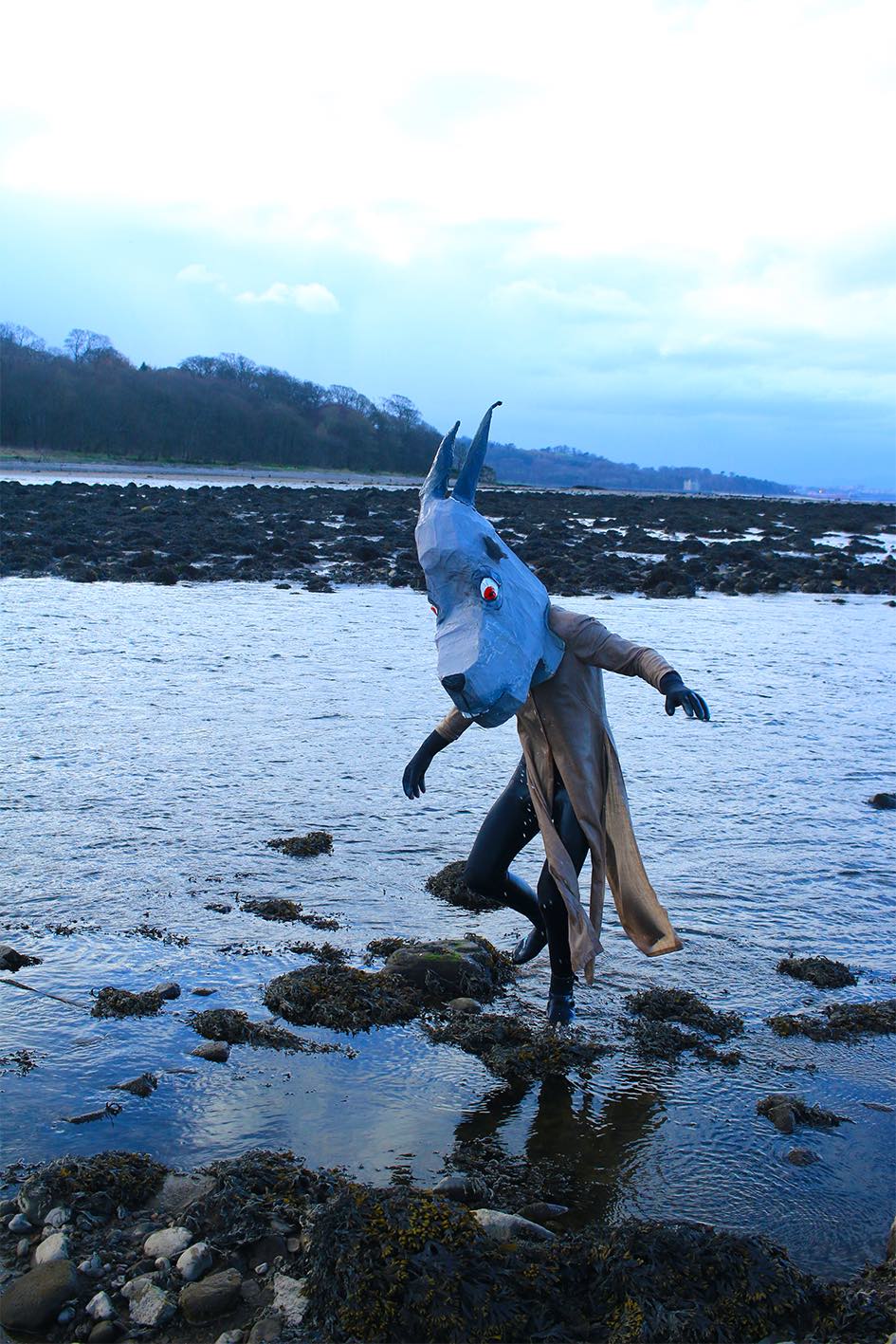 A figure wearing a papier-mache goats mask emerging from the water at the beach.