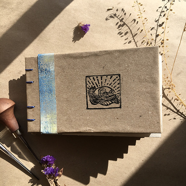 A photo of an A5 hand bound hard back portfolio book. The book is light brown with my logo lino printed onto the front (a square logo of the sea and sun) and bound using blue thread.