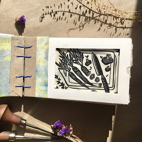 A photo of my A5 hand bound hard back portfolio book that contains samples of work from my A6 lino print series. The book lies open showing an example page, each page is a small envelope containing a removable print, with a window cut out to preview the work. Work features is Carrots and Thyme. 