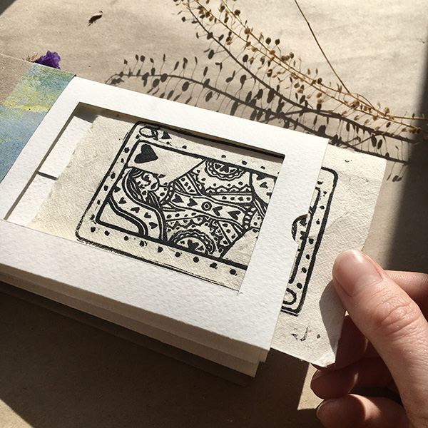 A photo of my A5 hand bound hard back portfolio book that contains samples of work from my A6 lino print series. The book lies open showing an example page, each page is a small envelope containing a removable print, with a window cut out to preview the work. Work features is Queen of Hearts- a print of a playing card.