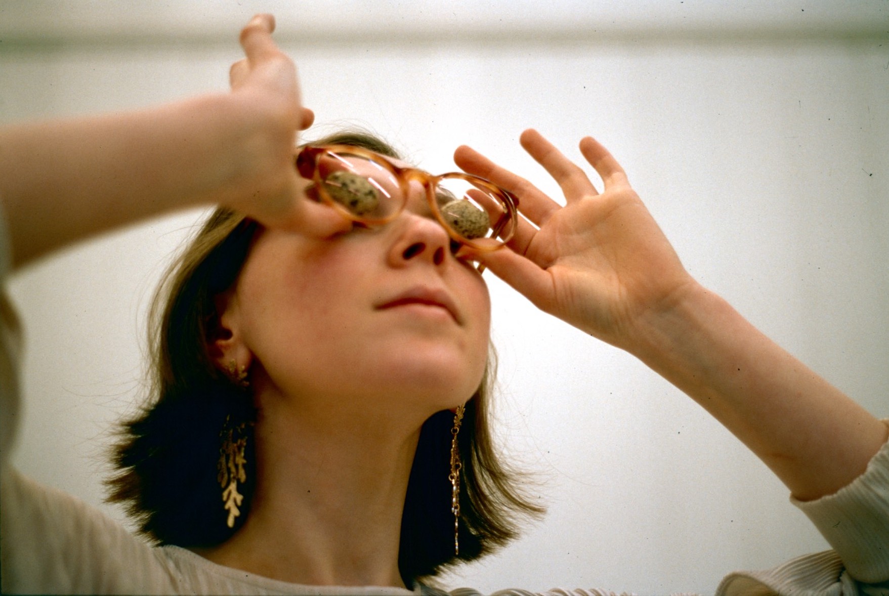a woman holds two quails eggs to her eyes and look out of the frame, as if to space