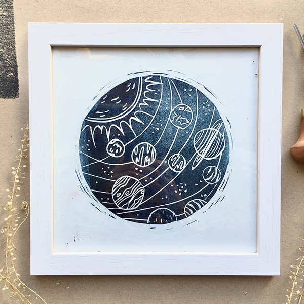 A circular lino print design of the solar system, printed in Prussian blue ink onto white paper. Framed in a square white wooden frame. 