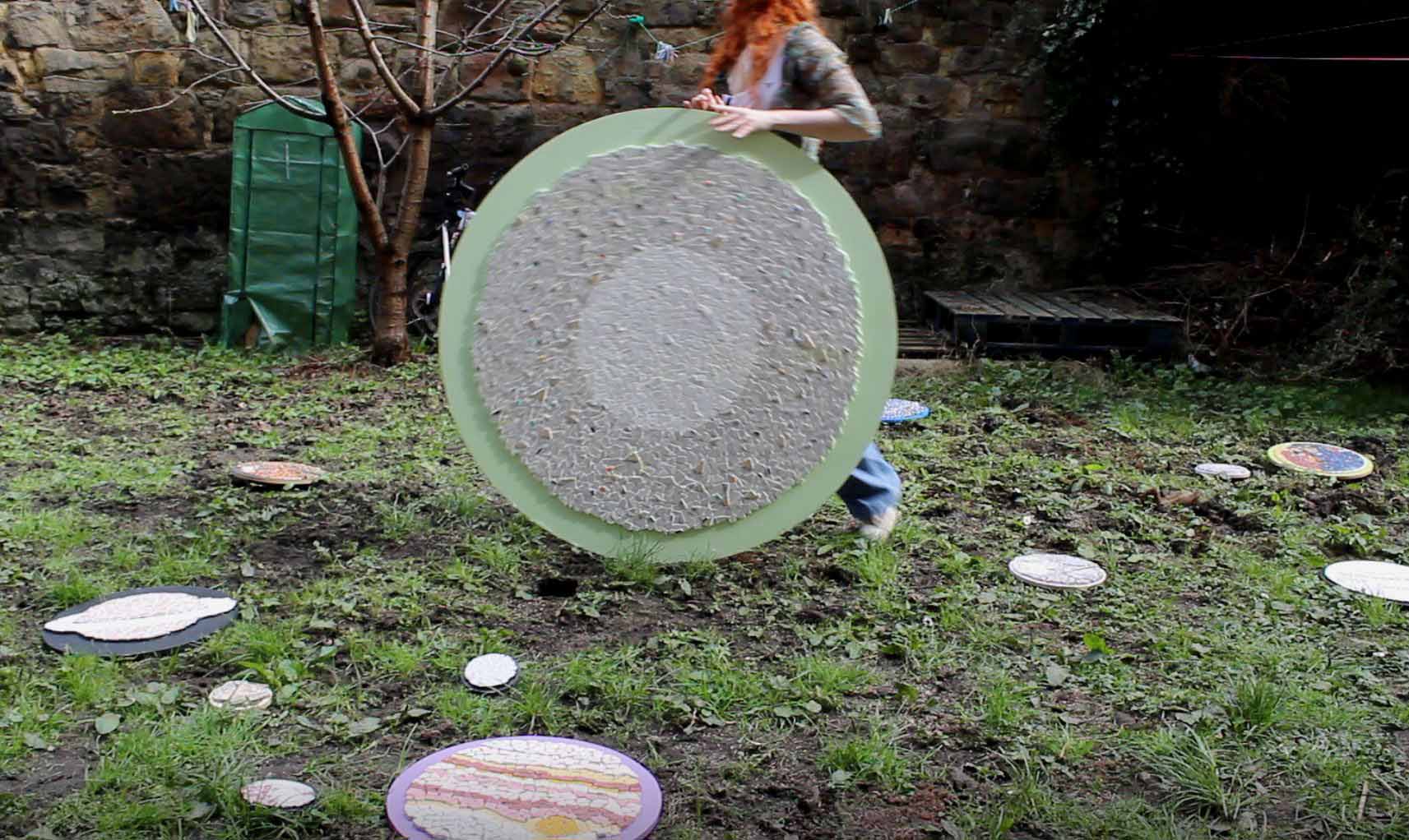 Large mosaic of the sun in grey, white and fractals of colour on green wood being rolled across grass. On the grass there are 12 other planets and astroids and moons. The person moving the sun has red hair
