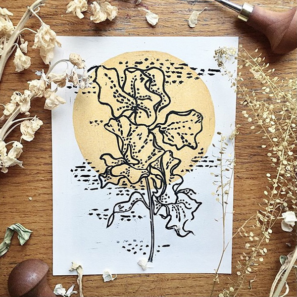 A lino print design in black ink of a detailed sweet pea flower surrounded by decorative dots, with a gold circle behind it to add a pop of colour. 