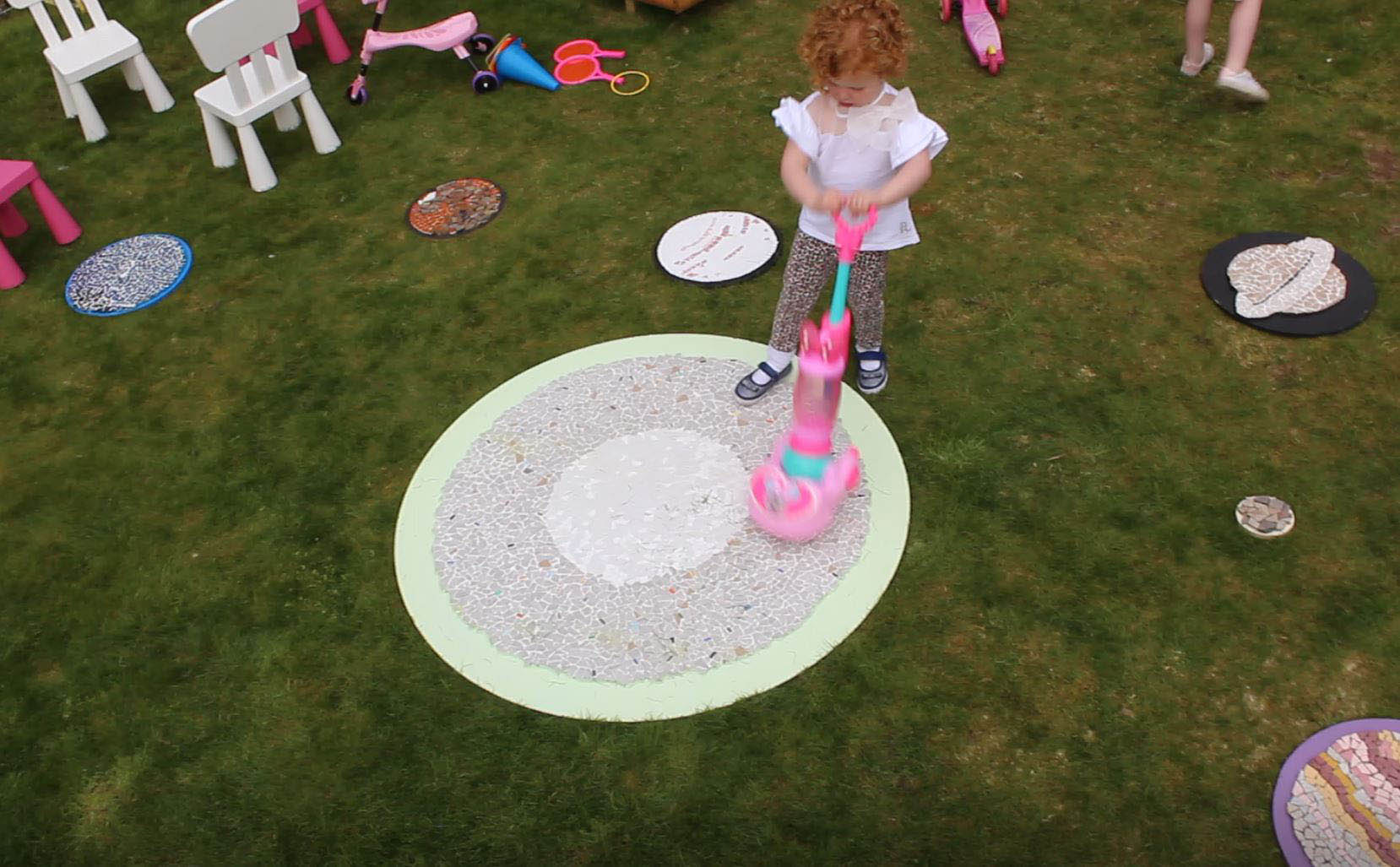Young girl pretending to hoover the sun mosaic with a pink childrens hoover. The mosaic is on grass and there are chairs and toys in the background