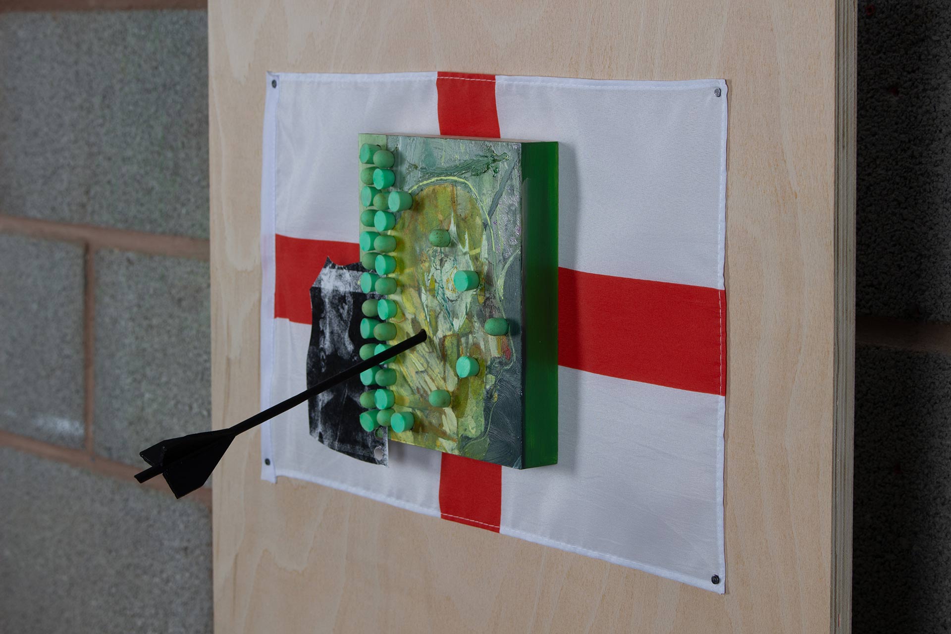 Little Englander, (2021) Oil and pencil on board embedded with wooden painted arrow, earplugs  and handmade photogram mounted onto England flag, 61.1 x 43.6 x 25 cm