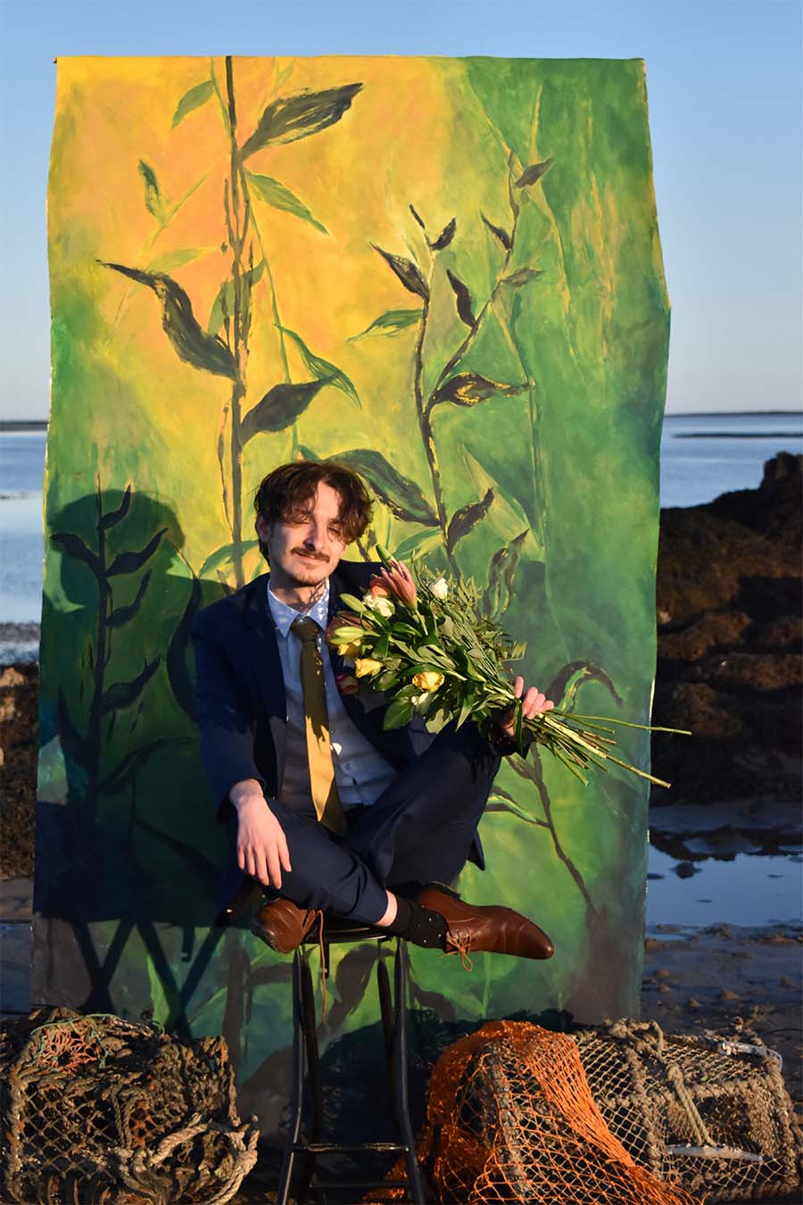 Model sits on a stool with his legs crossed, holding a bouquet of flowers and smiling. Behind him is a yellow and green painted backdrop of seaweed. At his feet are fishing nets.