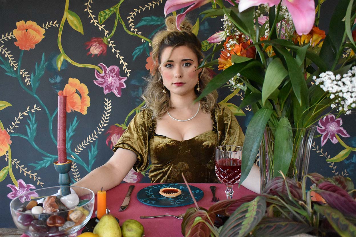 Actress stares into the camera, in front of her is a plate with a jam tart on it. The table is lavishly set with candles and flowers, and the backdrop is a painted wallpaper with a floral pattern