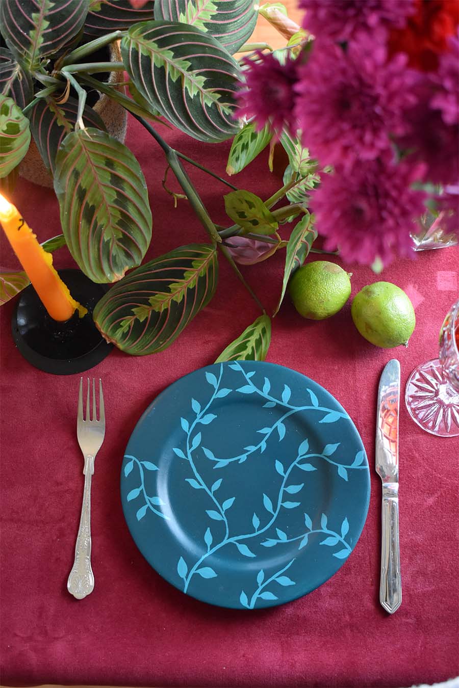 A table setting featuring a blue painted plate with vines on a red velvet tablecloth 