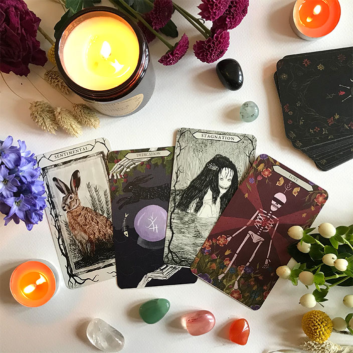 Illustrated tarot cards are spread over a table, surrounded with gemstones and candles