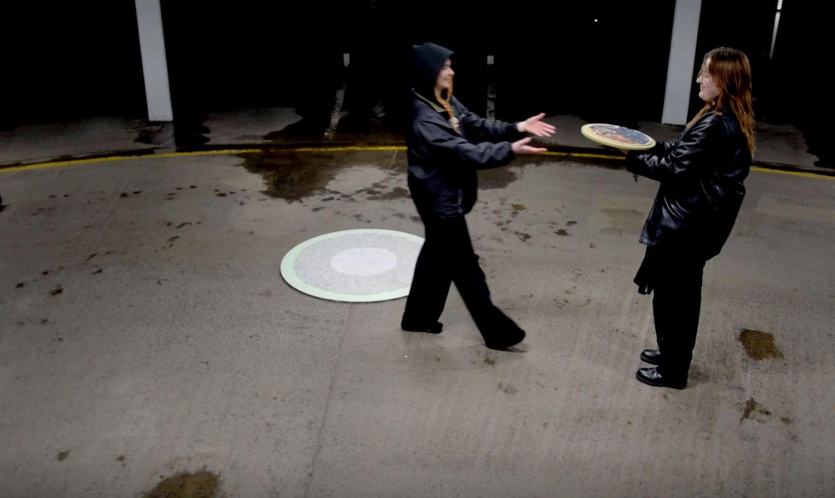 Inside a parking lot a woman is giving the artist a mosaic of the Earth. In the background is a mosaic of the sun. Both partcipants are wearing all black clothing and standing on grey gravel