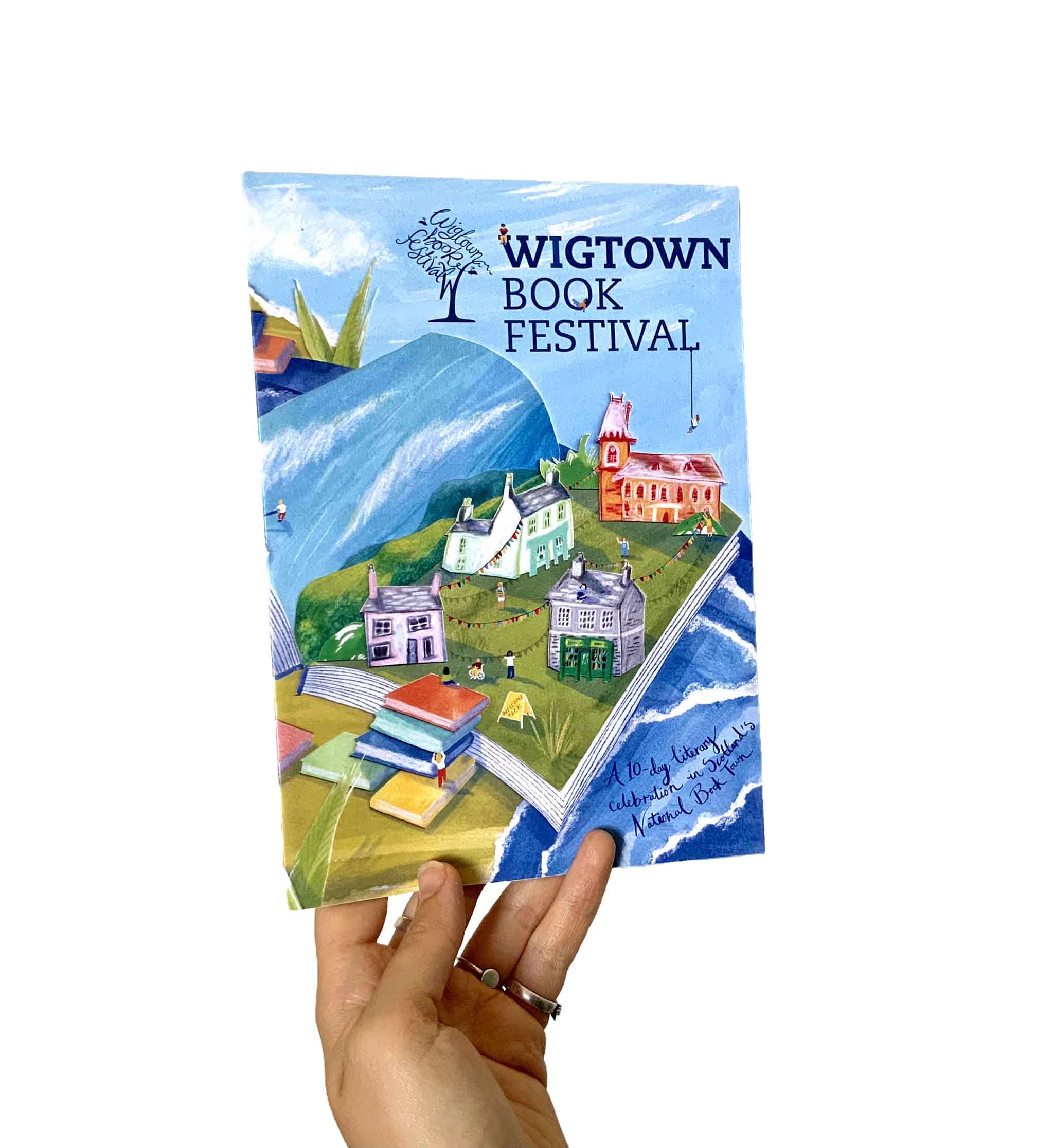A hand holds up the cover of a book with the title 'Wigtown Book Festival'. The scene is green and blue and features a large open book with small houses. 