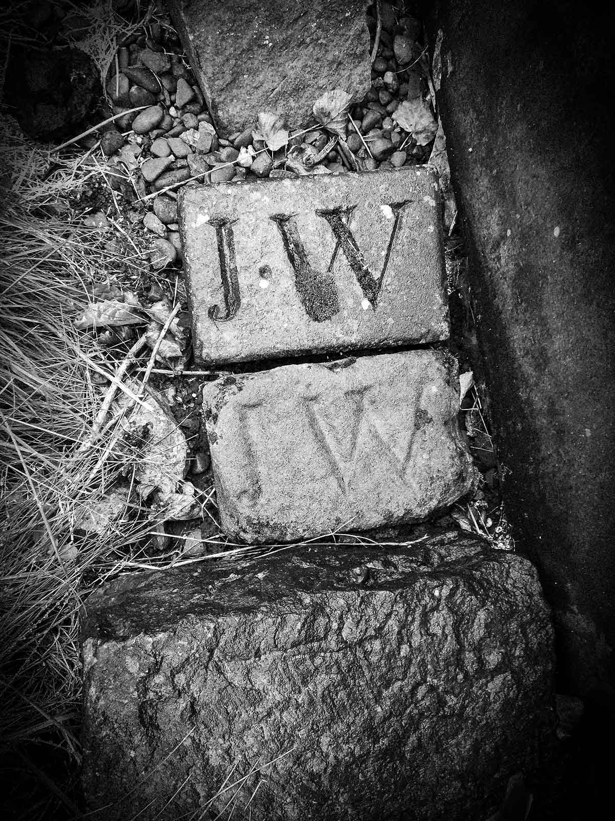 Greyscale analogue photograph of two bricks embeded in dirt soil, both with serift text J.W.