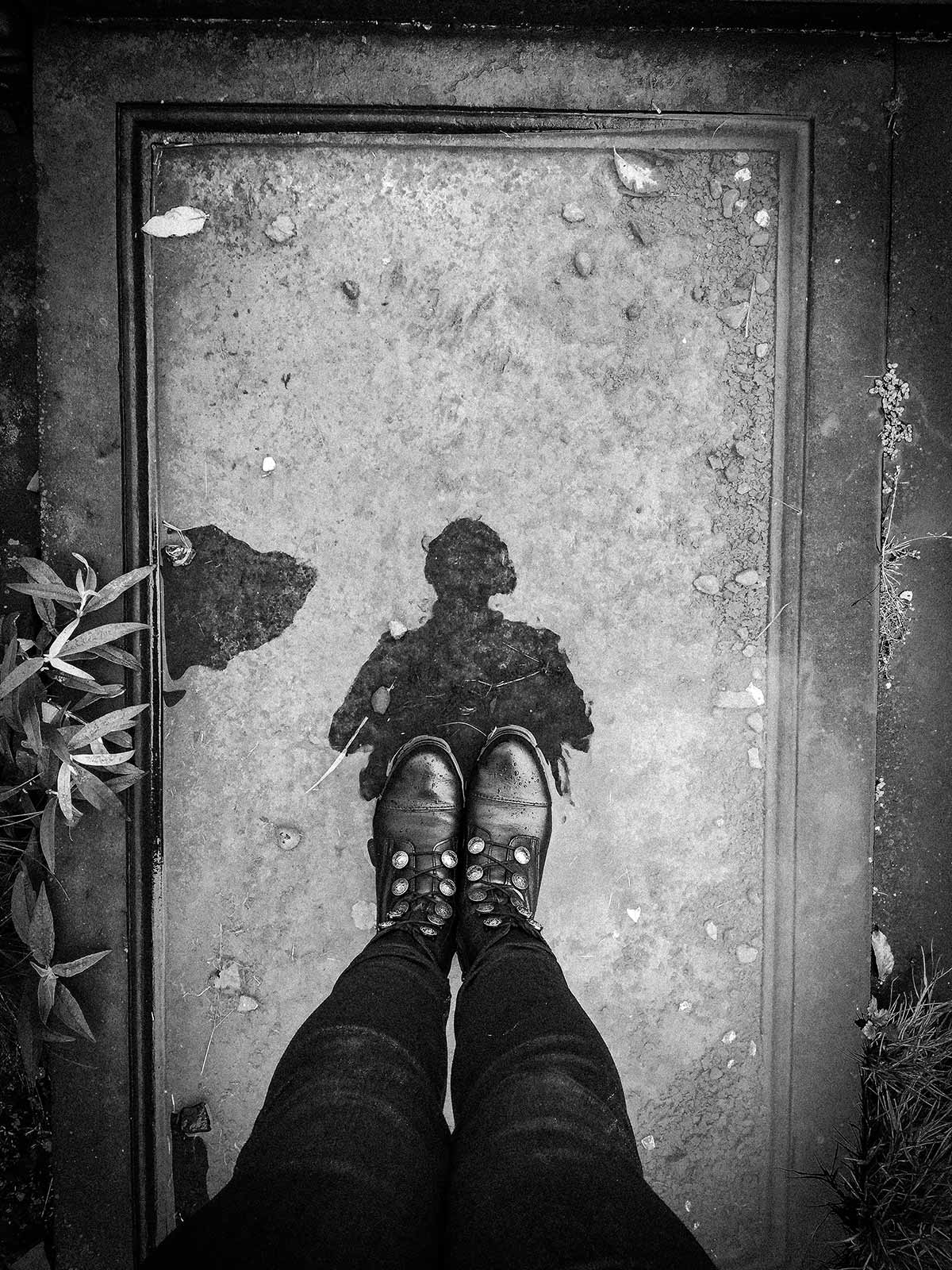Greyscale analogue photograph of the photographer's feet standing on a fallen headstone covered in water, the reflection of the photographer's outline visible in the water.