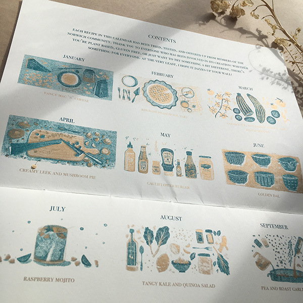 A photo of my illustrated 2022 Seasonal recipe calendar. It lays open showing the contents page with the illustration and recipe for each month. Risoprinted onto white paper in teal and gold. 