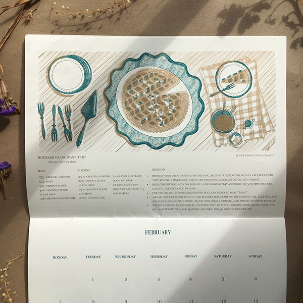 A photo of my illustrated 2022 Seasonal recipe calendar. It lays open showing my illustration for February, a rhubarb tart on a patterned plate with a slice cut out. Beside it are a pile of plates, cake forks, and a slice of tart on a plate. 6 patterned bowls with a gold textured background. Risoprinted onto white paper in teal and gold. 
