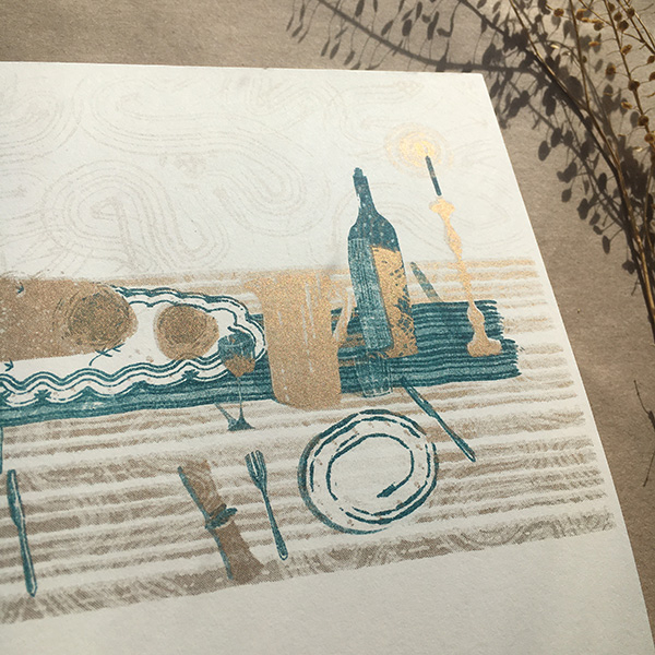 A photo of my illustrated 2022 Seasonal recipe calendar. Gives a close up look at my illustration of a mushroom wellington in the centre of a laid table, and a bottle of wine and candles. Risoprinted onto white paper in teal and gold. 