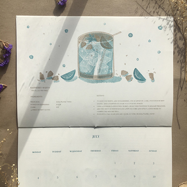 A photo of my illustrated 2022 Seasonal recipe calendar. It lays open showing my illustration for July- a cocktail glass of raspberry mojito and raspberries and lime around it. Risoprinted onto white paper in teal and gold. 