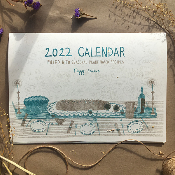 A photo of my illustrated 2022 Seasonal recipe calendar. It lays closed showing the front cover with my illustration for December- a mushroom wellington. Risoprinted onto white paper in teal and gold. 