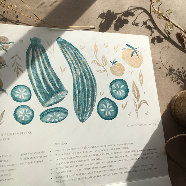 A photo of my illustrated 2022 Seasonal recipe calendar. It lays open showing my illustration for April, ingredients for savoury muffins including courgettes, tomatoes and herbs. 6 patterned bowls with a gold textured background. Risoprinted onto white paper in teal and gold. 