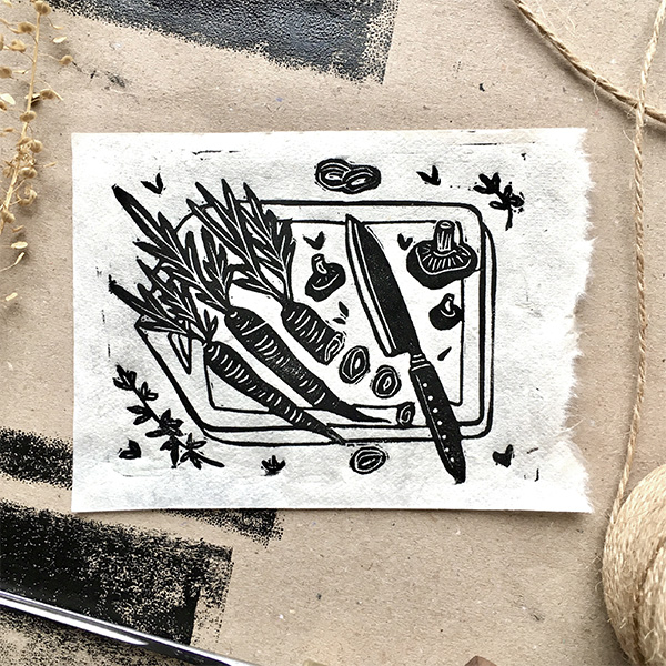 A lino printed design of carrots, mushrooms and sprigs of thyme on top of a chopping board and a knife laying next to a half chopped carrot. Printed in black on white paper. 