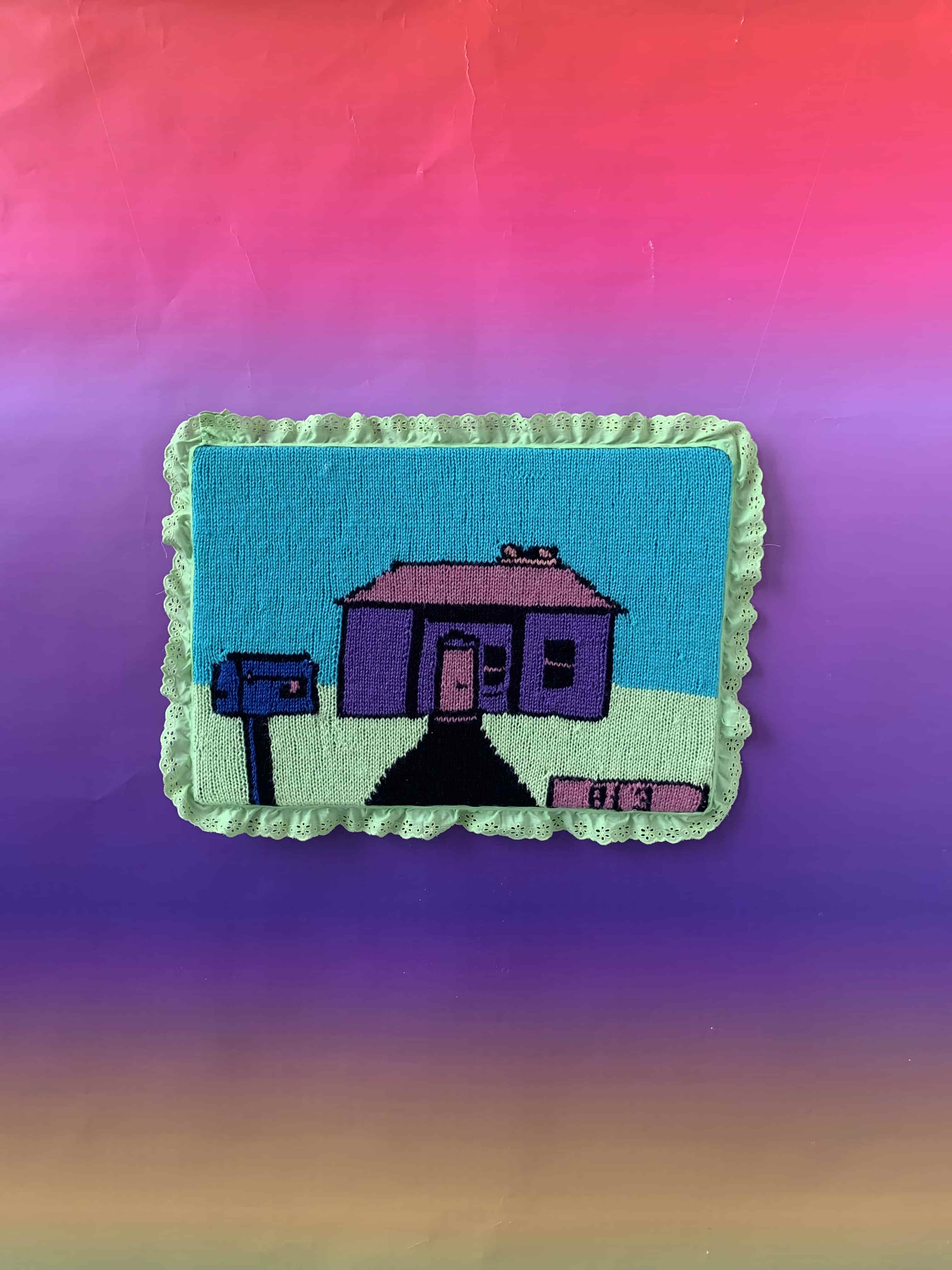 A knitted tapestry surrounded by green ribbon. It depicts a small pink and purple house with a purple letterbox.