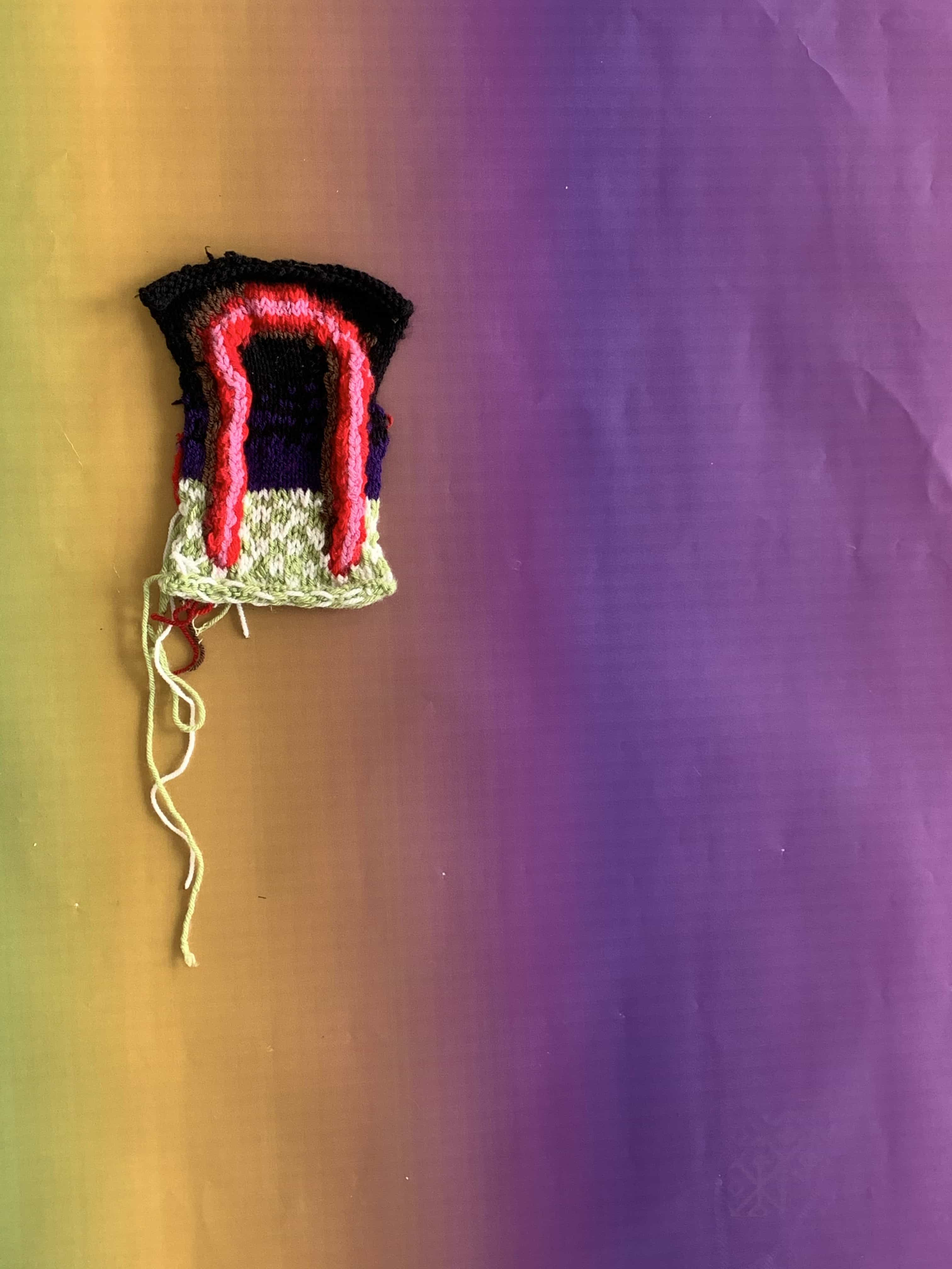 A small knitted tapestry on a gradient background. A black background, a red arch above a green and white checked bachground.