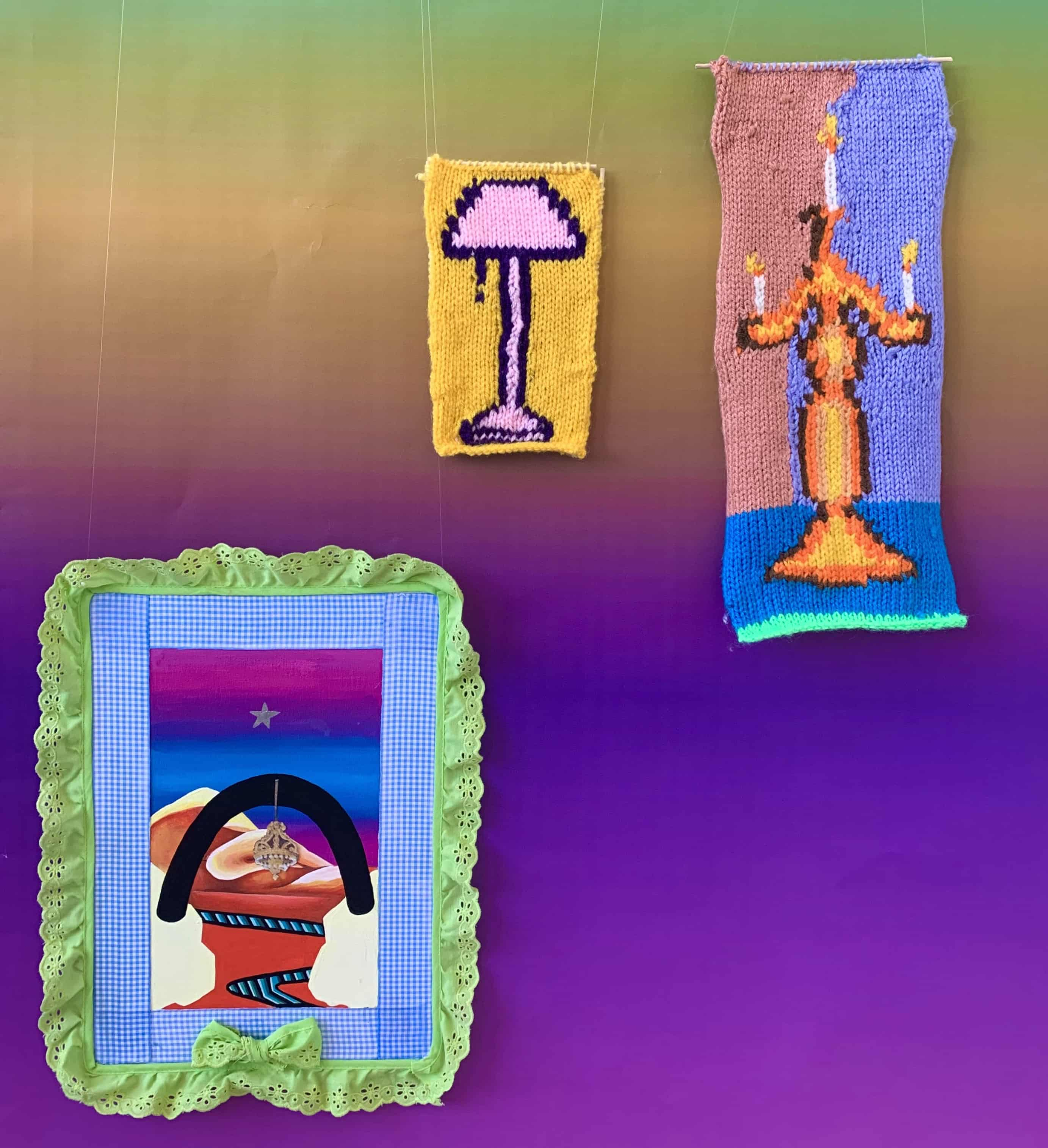 Three separate pieces hanging on a gradient background. One is a mixed media painting, and the other two are small knitted tapestries. The one on the right depicts an orange and yellow candelabra on a pink and purple background. The centre tapestry is a pink lamp on a yellow background. The painting on the left is a mixed media gouache painting surrounded by green and blue ribbon. In the centre are desert rocks , between which is a striped green and black river. A black arch hangs over the two front rocks.