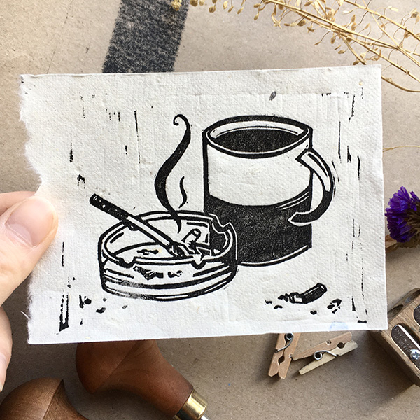 A lino print design of a mug of coffee and a cigarette in an ash tray with smoke whisping upwards. Printed in black on white paper.