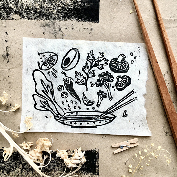 A lino print design of a bowl, chop sticks, and ingredients for ramen including broccoli, mushrooms, lime, chilli, garlic, pad choi etc. Printed in black on white paper.