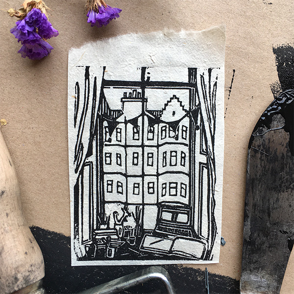 A lino printed design of a desk at a window, with a laptop, mug of pencils and an open sketch book on the desk, and bunting and curtains framing the window. The view looks out onto tenement Edinburgh buildings. Printed in black ink on white paper. 