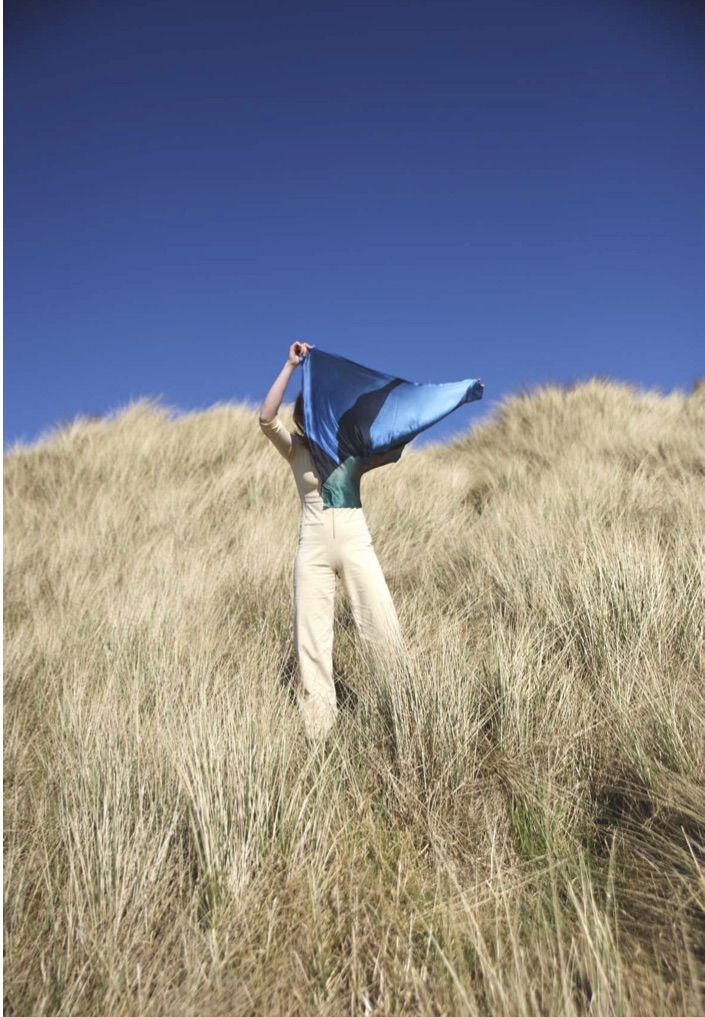 Figure standing in long sea grass holding silk textiles blowing in the wind.
