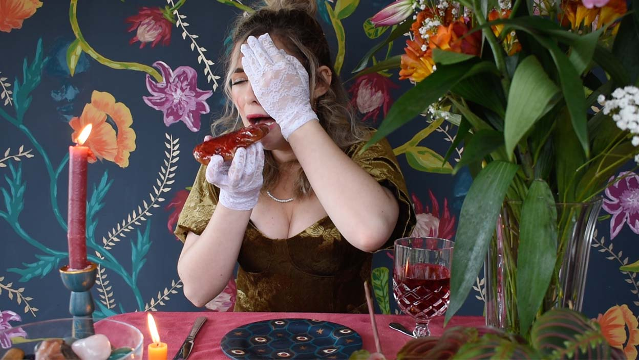 Actress holds a hand over her eye and is eating a heart with her other hand with a pained expression on her face. 