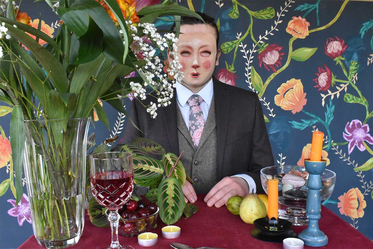 A masked man in a suit stares into the camera. He is sitting at a table lavishly set with candles and flowers. The backdrop is a painted wallpaper with a floral pattern