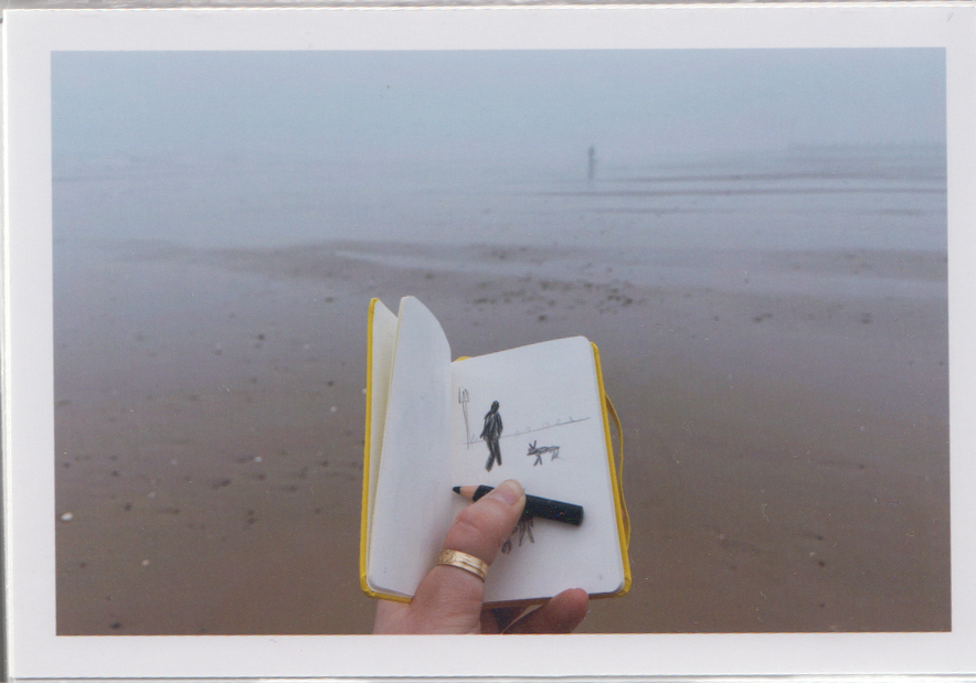 a misty beach in winter, a hand holds a yellow sketchbook with silhouettes on the pages