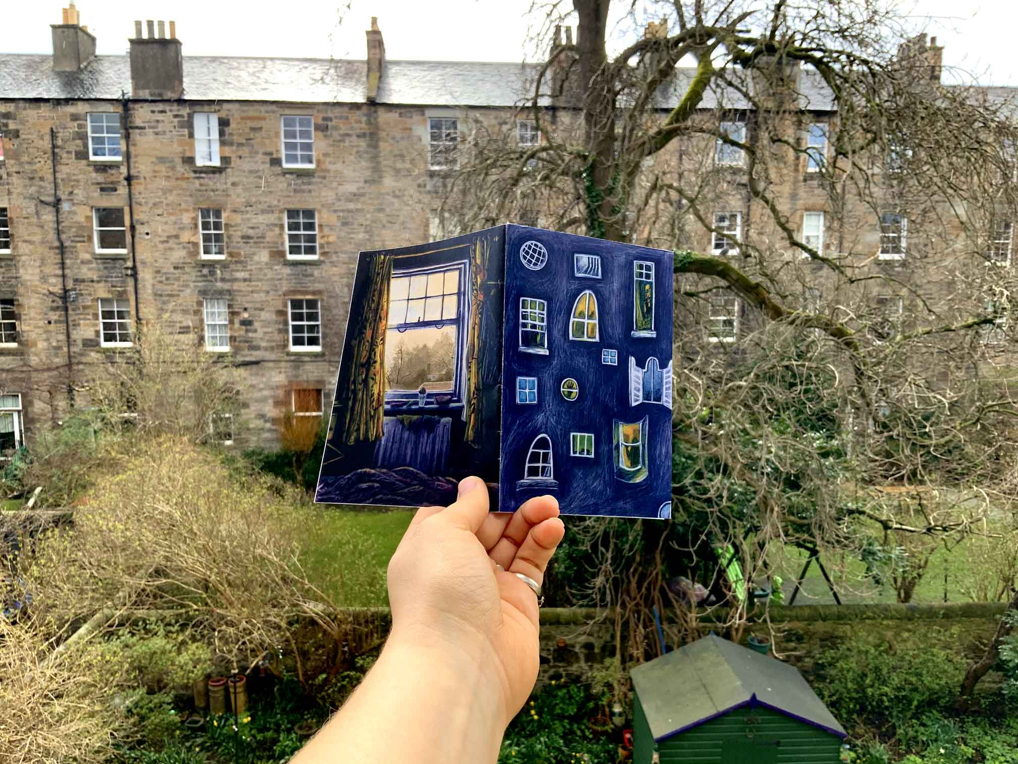 A photo of a hand holding up a small Zine with a picture of windows on