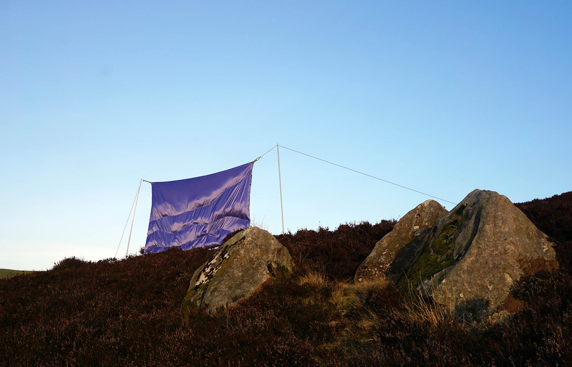 Lilac Taffeta stuffed sculpture held up by two poles on top of the topography of a Moore of heather in the fore grown are three large stones 
