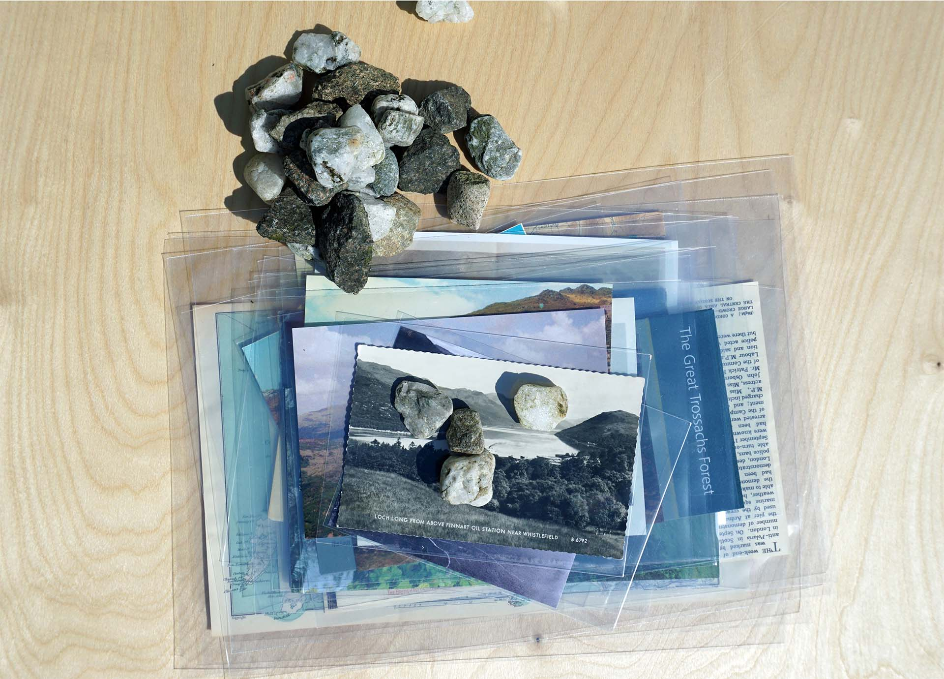 A view of collected archival materials layered on top of each other held down by a pile of stones on a birch ply table. 
