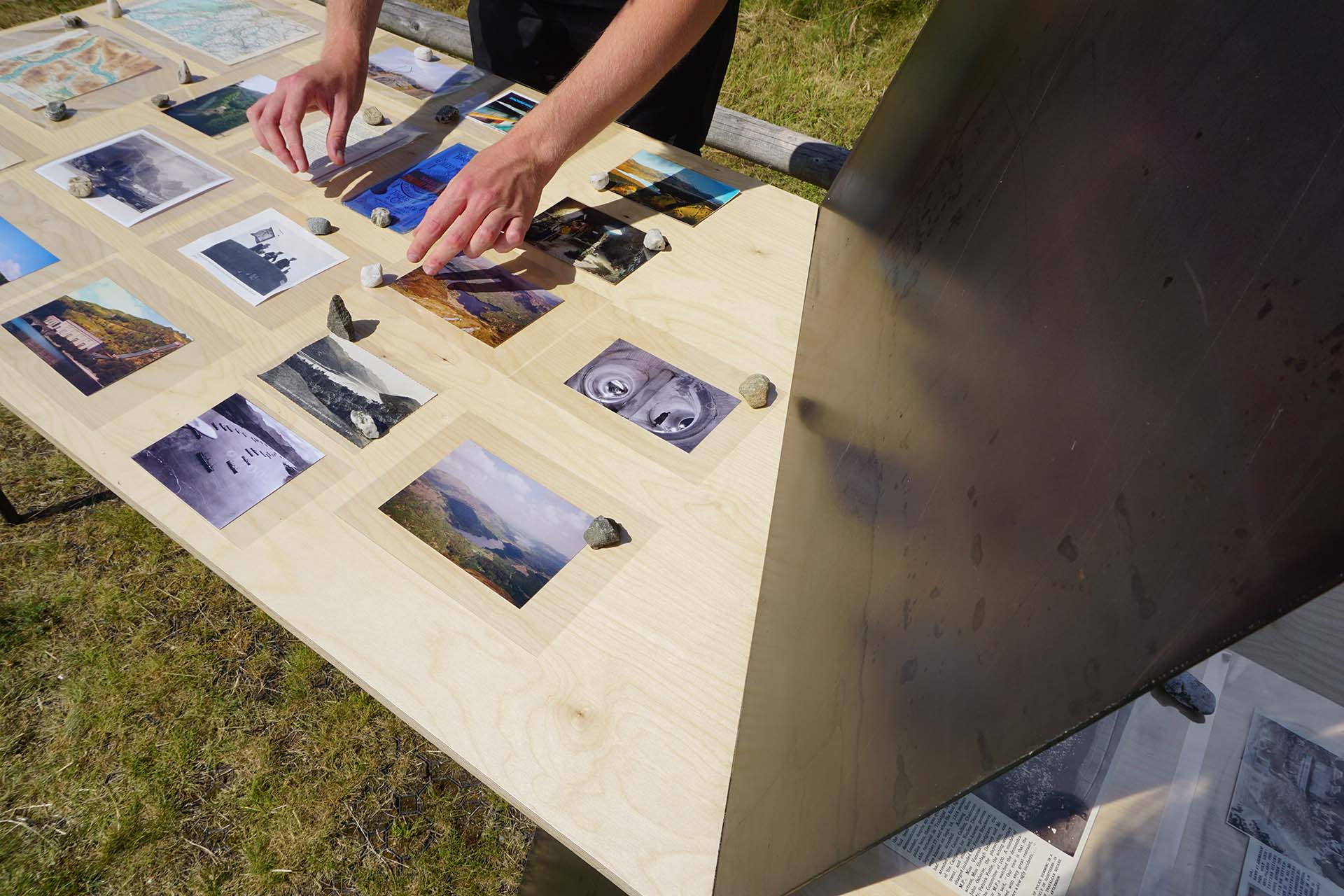 Collected archival materials displayed on birch ply table with hands reaching down to view an archival map of the Trossachs, these hands are foggly reflected in the dark hot rolled steel. 