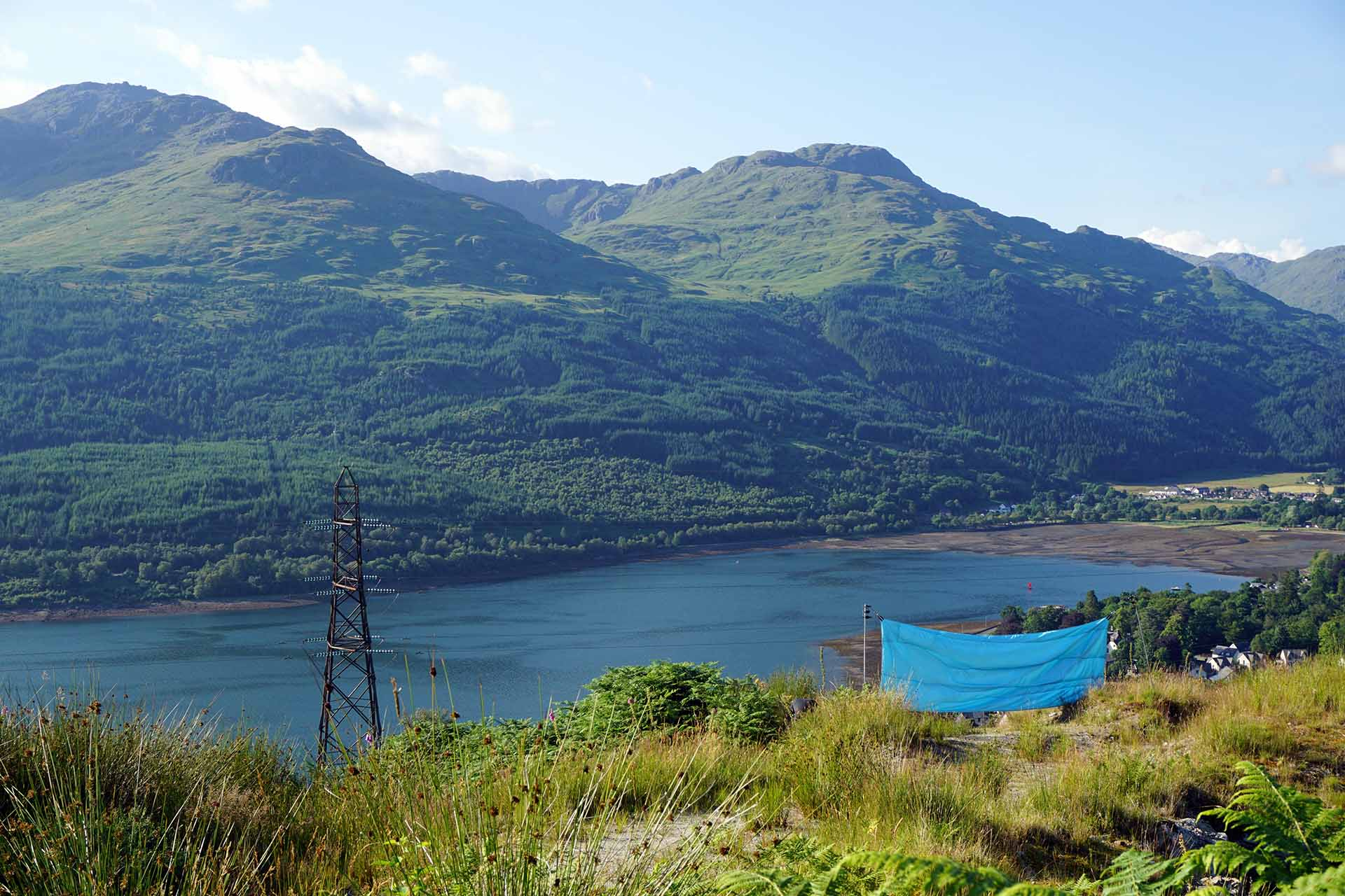a faraway view of baby blue two toned taffeta (1.5 m x 4.5 m) stretch across a path situated half way up a mountain. The blue fabric looks over loch long and out towards more mountains. 