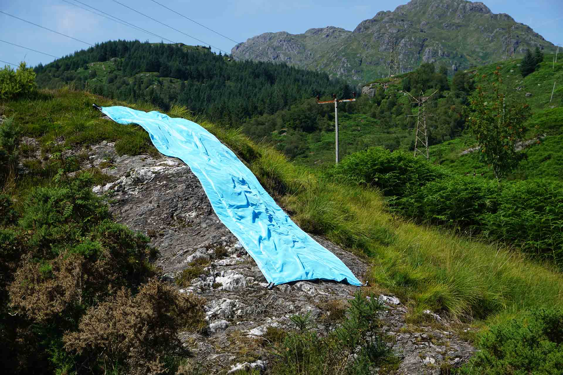 Baby Blue two toned taffeta stuffed with pattern sewn onto it draped over the topography of Arrochar. The fabric cascades down a large grey rock face and looks like water