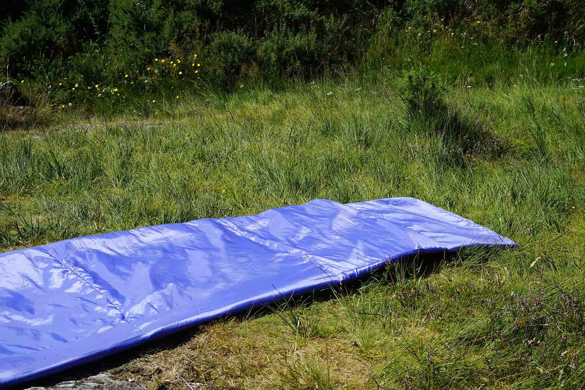 Lilac two toned stuffed taffeta in the shape of a sleeping bag wind sock hybrid two tone lilac soft sculpture placed on the ground that is a vivid shade of green due to the long summer grass